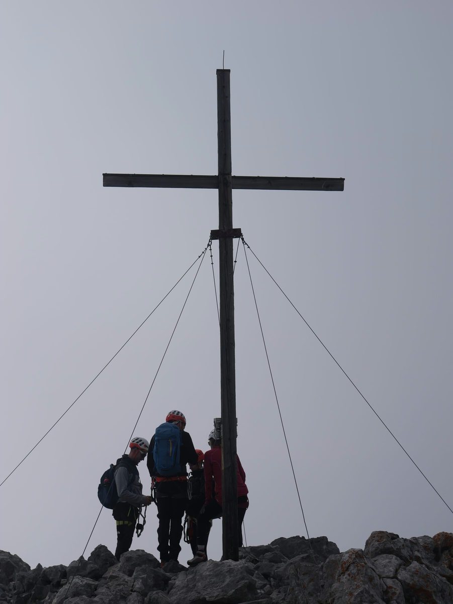 Hohes Gipfelkreuz als Silhouette vor grauem Himmel, darunter eine kleine Gruppe Bergsteiger