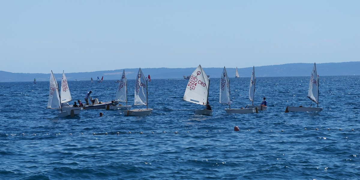 Kinder in Optimist-Jollen segeln auf tiefblauem Meer, dahinter eine Insel